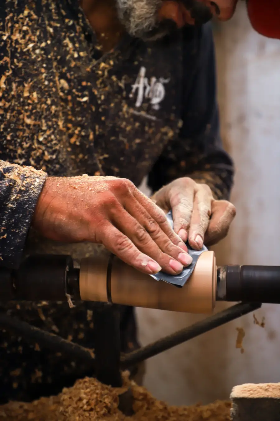 An artisan’s hands sanding a turned olive wood piece held on the lathe, shavings covering his apron and the bench below